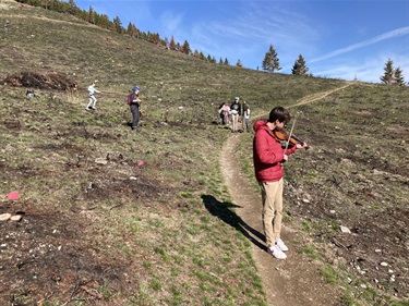 A violinist plays music on a trail while students plant trees.