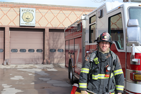 Firefighter standing in front of an engine and fire station.